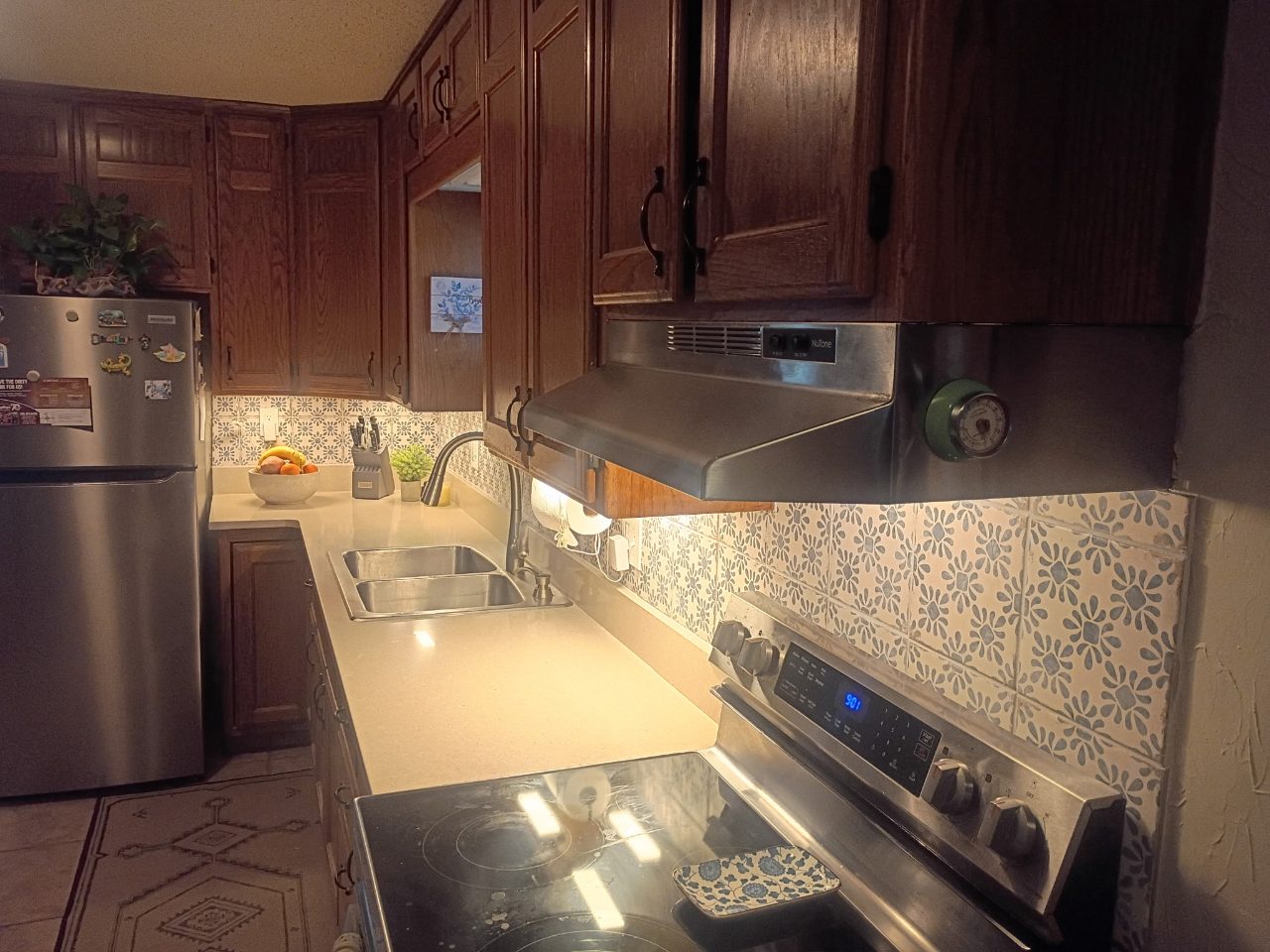 Blue and white patterned tile backsplash behind a kitchen sink and range, lit by under-cabinet lights